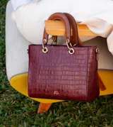 Cartera de mano de estructura rígida con textura símil cocodrilo en color bordó, con manijas superiores cortas en tono tostado y herrajes metálicos dorados.