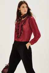 Camisa de manga larga en color negro con tejido semitransparente y detalles de hilos metalizados. Presenta un diseño con bordados florales en tonos tierra, cierre frontal con botones y un lazo decorativo en el cuello.