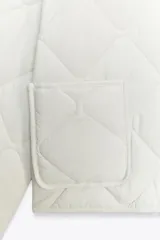 Chaqueta acolchada de cuello redondo y manga larga, con cierre frontal de botones y dos bolsillos delanteros de plastrón. Presenta un diseño de costuras en forma de rombos.