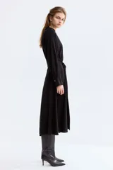 Vestido camisero midi de color negro, corte holgado con cuello, escote en V y cierre frontal de botones. Cuenta con mangas largas con puños anchos y un cinturón ajustable para anudar en la cintura.