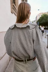 Blusa de manga larga con estampado de cuadros vichy en blanco y negro. Presenta un escote en V adornado con un volante amplio y un lazo decorativo en el centro. Las mangas terminan en puños elásticos.