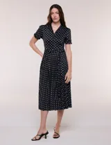 Vestido midi negro con estampado de lunares blancos, de la marca Sharagano. Presenta cuello con solapa, escote en V, cierre frontal con botones y mangas cortas. Tiene un corte en la cintura con lazo ajustable y falda plisada.