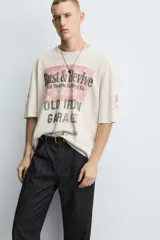 Camiseta de punto de algodón con corte boxy fit, cuello redondo y manga corta. Presenta un estampado frontal con temática de taller mecánico en tonos rojo desgastado y negro, incluyendo el texto "Rust & Revive", "Old Iron Garage" y detalles técnicos. La prenda tiene un efecto lavado y acabados irregulares.