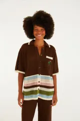 Camisa de punto color marrón con cuello solapa, cierre frontal de botones y mangas cortas. Presenta un bolsillo en el pecho con detalle bordado de una palmera y un tucán, y franjas horizontales multicolor en la parte inferior.