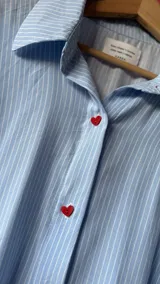 Camisa oversize de fibrana a rayas celestes y blancas, con cuello clásico, mangas largas con puños y botones frontales de acrílico en forma de corazón rojo.