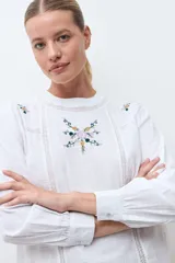 Blusa blanca de manga larga con cuello redondo, confeccionada en tela liviana con detalles de encaje vertical y bordado floral en el pecho.