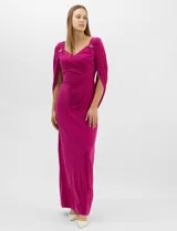 Vestido largo de seda fría color fucsia, con escote cruzado drapeado y apliques de pedrería en los hombros. Presenta mangas largas abiertas estilo capa y un drapeado lateral en la cintura que estiliza la silueta.