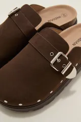 Suecos negros de estilo zueco con capellada de cuero sintético, detalle de hebilla metálica dorada y tachas decorativas alrededor de la suela. Poseen plantilla ergonómica y suela de plataforma dentada.