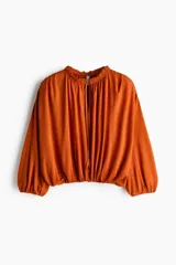 Remera corta oversize de viscosa en color terracota, con diseño drapeado, cuello alto con volantes y aberturas con tirantes finos en el frente. Cuenta con mangas tres cuartos abullonadas y elástico en los puños y bajo.