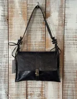 Cartera tipo morral de cuero color azul oscuro, con solapa frontal y cierre metálico de broche. Cuenta con asa de hombro ajustable con detalles de nudos y tiras decorativas en los laterales.