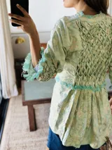 Camisa larga tipo sobrecamisa o abrigo ligero, color verde menta pálido con estampado floral en tono amarillo claro. Presenta escote en V con atado frontal mediante lazos, mangas largas con detalle fruncido y puños con flecos en tono turquesa.