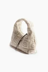 Cartera de mano tejida en crochet con motivo de cuadrados, color crudo, con asa corta integrada y tiras de cuero marrón como detalle de cierre.