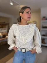 Blusa de tela liviana color crema con estampado floral pequeño en tonos rojizos. Presenta un diseño con volados en el frente y hombros, mangas largas abullonadas con puños elásticos y cuello redondo.