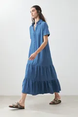 Maxi vestido camisero de jean azul con cuello clásico, manga corta, cierre frontal con botones y falda amplia con volantes.