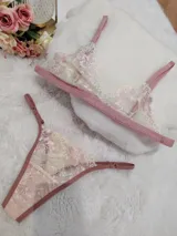 Conjunto de lencería compuesto por bralette y tanga regulable, confeccionado en encaje color champagne con detalles florales bordados en hilo rosado y terminaciones en cinta color rosa viejo.