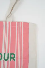Bolso tote de tela con estampado de rayas verticales rosadas y blancas, laterales y asas en color beige. Presenta la palabra "BONJOUR" en verde en el centro y "INDIAN" en la parte inferior.