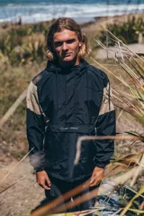 Buzo rompevientos tipo anorak con capucha y cordones ajustables. Presenta un diseño bicolor en negro y beige, con bolsillo frontal tipo canguro y logo de la marca Glassy Waves bordado en el centro. Posee puños elásticos y cierre frontal parcial.