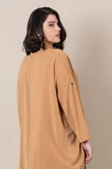 Camisa larga color camel, de corte irregular, más larga en la parte trasera, con botones dorados, manga larga con presilla para sujetar.
