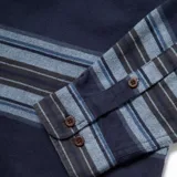 Camisa de franela a rayas horizontales en tonos azul marino, celeste y gris. Tiene cuello clásico, cierre frontal con botones, dos bolsillos de parche con botón en el pecho y mangas largas con puños abotonados.