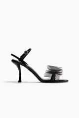 Sandalias de taco alto negro con acabado charolado, tira delantera con detalle de lazo voluminoso de tul y tira ajustable al tobillo con hebilla metálica.