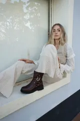 Bota de caña media confeccionada en cuero color marrón oscuro, con suela track negra de goma. Presenta un diseño estilo western con correas decorativas, remaches metálicos y argollas plateadas en el lateral.