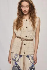 Chaleco de punto color beige, sin mangas, con escote redondo y cierre frontal con botones carey. Presenta diseño calado y silueta amplia.