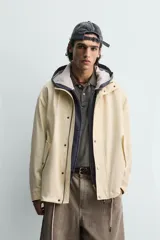 Parka de tejido técnico color beige claro, corte relajado (relaxed fit), con cuello subido, cierre frontal oculto por solapa con botones a presión y bolsillos de solapa en cadera.