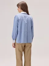 Camisa de manga larga en color azul con textura tipo denim, con cuello y puños en contraste de color blanco. Presenta costuras decorativas horizontales y verticales en el frente, cierre frontal con botones y corte recto.