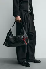 Bolso tipo bandolera de cuero sintético negro con acabado brillante. Presenta un compartimento principal con cierre de cremallera, un bolsillo frontal con cierre y detalle de charms colgantes decorativos. Incluye asa de hombro ajustable.