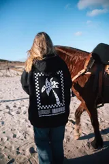 Hoodie oversize negro con capucha, con gran estampado blanco en la espalda que presenta un diseño de palmera dentro de un marco a cuadros y texto.