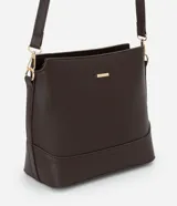 Cartera tipo hobo de tamaño pequeño, confeccionada en material sintético color marrón oscuro con acabado liso. Presenta una correa ajustable, cierre principal con cremallera y detalles metálicos dorados, incluyendo una placa con el logo de la marca en el frente.