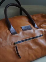 Bolso tipo weekender de cuero marrón, con asas de mano y bolsillo frontal con cierre metálico.