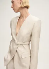Blazer de lino color beige con diseño cruzado, cuello de pico y hombros estructurados. Incluye un cinturón ajustable con hebilla del mismo tejido y dos bolsillos laterales con solapa.