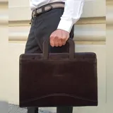Sobre ejecutivo tamaño oficio de diseño rígido y rectangular. Confeccionado en símil cuero color marrón oscuro con textura veteada. Cuenta con cierre perimetral, manijas integradas y logo de la marca grabado en el frente.