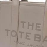 Bolso tipo tote de gran tamaño, confeccionado en cuero marrón con textura granulada. Presenta el logo de la marca estampado en el frente con la inscripción The Tote Bag Marc Jacobs. Cuenta con asas superiores fijas y un diseño estructurado y espacioso.