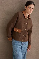 Campera corta de paño color marrón con detalles en cuero sintético al tono en cuello, puños y solapas de bolsillos. Presenta cierre frontal con botones metálicos dorados y dos bolsillos frontales tipo cargo.