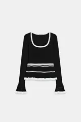 Jersey de punto acanalado en color negro con detalles en blanco. Presenta escote redondo, mangas largas con puños acampanados y detalle de volante en el bajo.