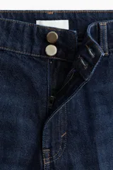 Jean de denim azul oscuro, de tiro alto y piernas amplias con pliegues frontales y traseros. Cuenta con cierre y doble botón metálico en la cintura.