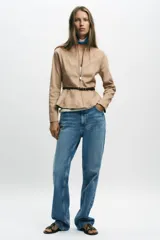 Chaqueta color beige con efecto ante, cuello redondo, manga larga y cierre frontal con cremallera. Presenta un bajo en evasé y un cinturón fino de cuero marrón oscuro con hebilla metálica.