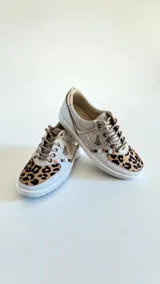 Sneakers de mujer en tonos blancos, dorados y detalles con estampado animal print de leopardo en la puntera. Incorporan cordones frontales y apliques metálicos decorativos.