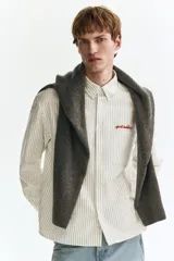 Camisa de corte holgado en mezcla de algodón Oxford, con diseño a rayas, cuello americano y cierre frontal de botones. Cuenta con un bolsillo superior en el pecho con texto bordado en rojo, mangas largas con puños abotonados y bajo recto.