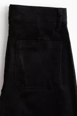 Pantalón de pana de algodón color negro, de tiro alto, con pliegues delanteros y traseros. Cierre con dos botones dorados, bolsillos traseros abiertos y piernas rectas.