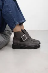 Botines de cuero vacuno gamuzado con textura envejecida, color gris oscuro, con suela track de goma dentada y detalle de gran hebilla metálica plateada en el lateral con solapa cruzada.