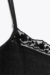 Vestido corto negro de gasa con escote recto y tirantes finos ajustables. El corpiño tiene fruncido elástico y el ruedo es escalonado con volados. Presenta un detalle de encaje a tono en el borde del escote.