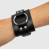 Brazalete ancho de cuero vegano negro, con una gran argolla metálica plateada al frente sujeta por tiras con remaches.