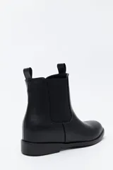 Bota tipo chelsea de caña media confeccionada en cuero sintético, con paneles laterales elásticos y presillas superiores. Presenta un diseño minimalista con suela plana y punta redondeada.