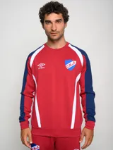 Buzo deportivo de manga larga, color principal rojo, con mangas azul marino y detalles en blanco que bajan desde el hombro hasta el puño. Presenta cuello redondo, logo Umbro y escudo del Club Nacional de Football en el pecho.