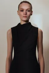 Vestido midi de color negro, con cuello subido y diseño sin mangas. Presenta un detalle de drapeado lateral en la parte delantera y cierre de cremallera oculta en la espalda.