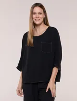 Blusa oversize color negro con caída fluida, escote redondo, bolsillo frontal en el pecho, mangas tres cuartos de hombro caído y costuras a la vista en contraste.