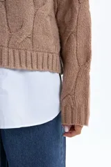 Sweater de punto en color beige con diseño de trenzas verticales, cuello alto acanalado y mangas largas con puños ajustados.